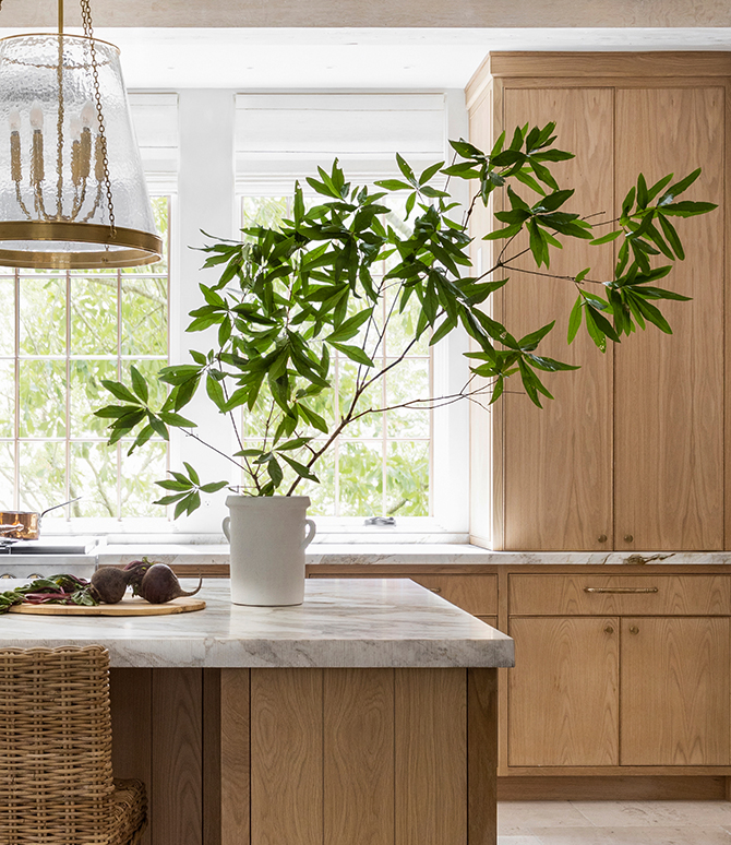 A bright kitchen with marble island, potted plant, wicker stools, wooden cabinets, and large windows.