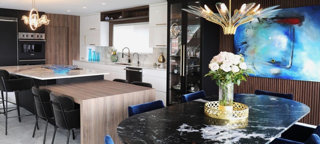 Modern kitchen with a combination island and dine-in table fixture adjacent to a dining area with a marble-topped table, velvet chairs, and a striking chandelier. 