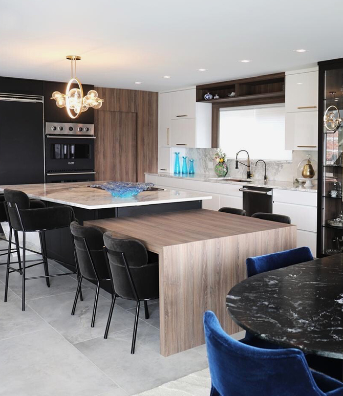Modern kitchen with a combination island and dine-in table fixture adjacent to a dining area with a marble-topped table, velvet chairs, and a striking chandelier. 