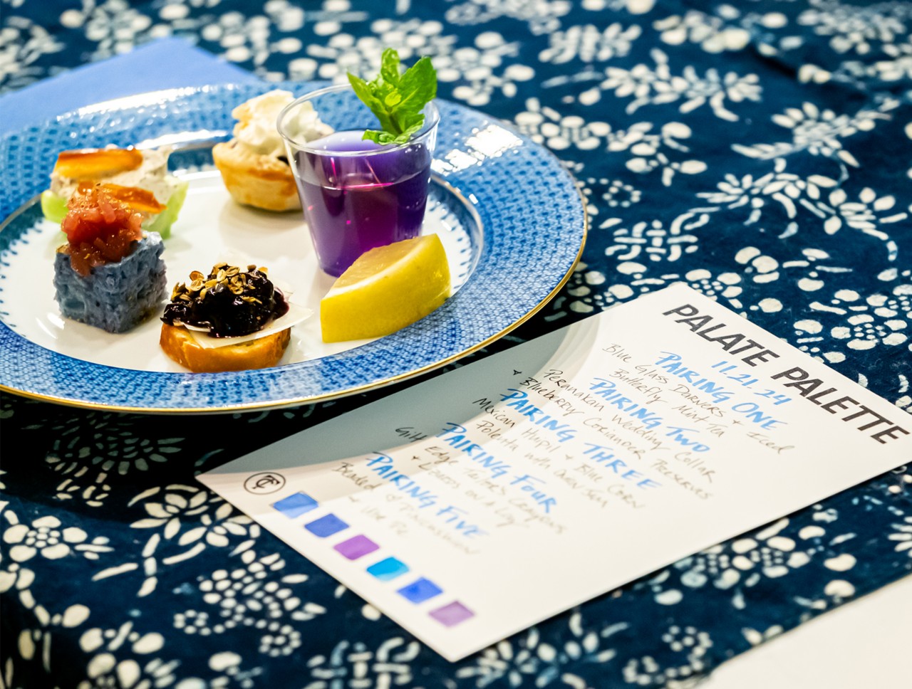 A plate with various appetizers inspired by the color blue and a menu card on a blue patterned tablecloth.