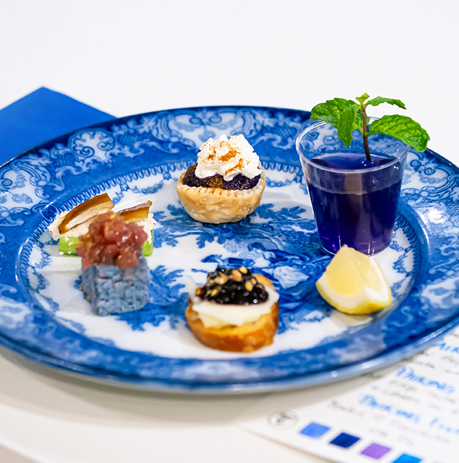 A blue patterned plate featuring small bites and a plastic cup of tea with a mint sprig and lemon wedge.