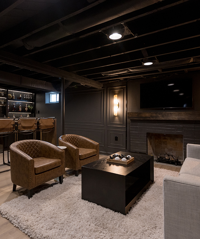 Basement room, color-drenched in Sherwin-Williams Iron Ore, with a home bar, brown leather stools, and a seating area with a sofa and armchairs.
