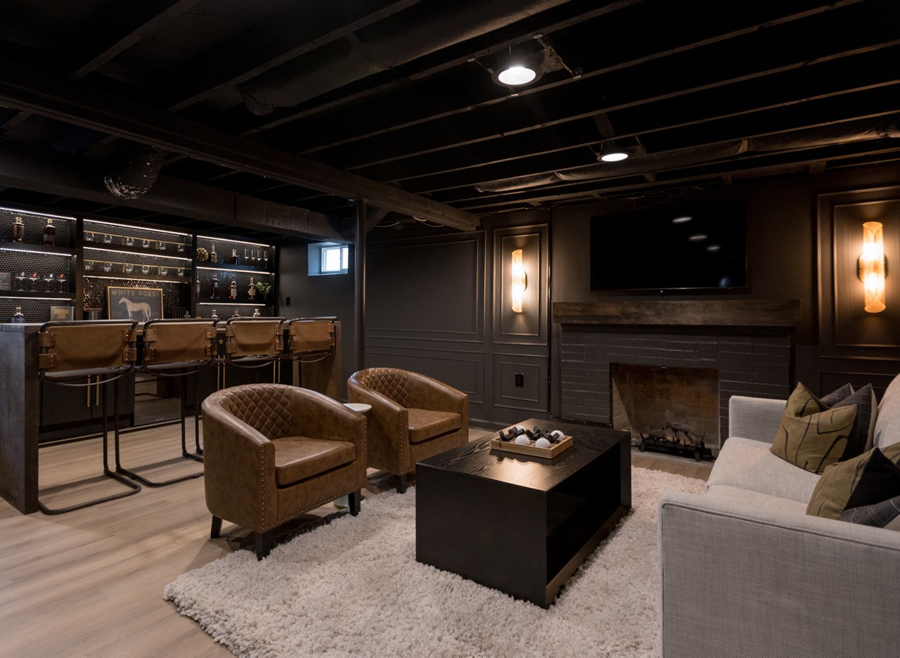 Basement room, color-drenched in Sherwin-Williams Iron Ore, with a home bar, brown leather stools, and a seating area with a sofa and armchairs.