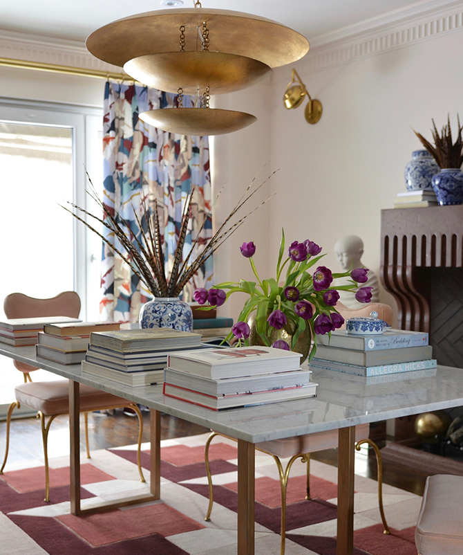A room painted Sherwin-Williams Faint Coral with a marble table topped by piles of books, vases of feathers and tulips, under a gold chandelier, near a fireplace with decorative items.