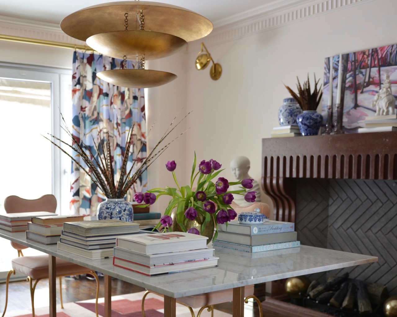 A room painted Sherwin-Williams Faint Coral with a marble table topped by piles of books, vases of feathers and tulips, under a gold chandelier, near a fireplace with decorative items.