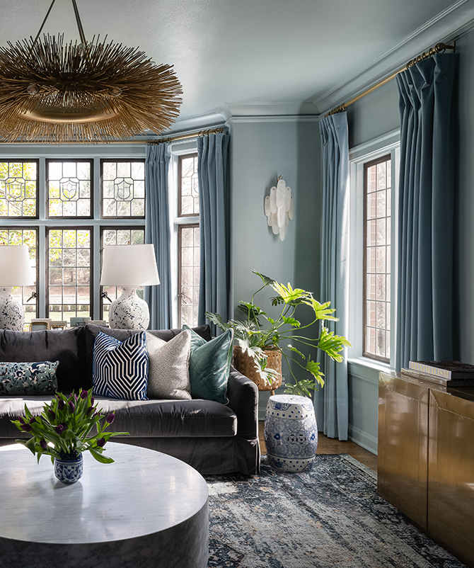 Living room with a dark gray sectional sofa, assorted pillows, marble coffee table with greenery, and tall windows with blue curtains.