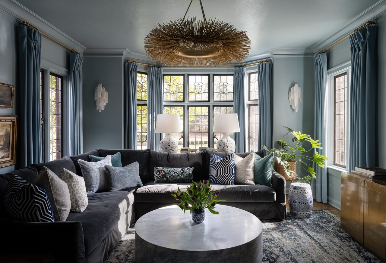 Living room with a dark gray sectional sofa, assorted pillows, marble coffee table with greenery, and tall windows with blue curtains.