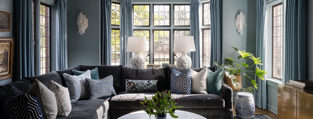 Living room with a dark gray sectional sofa, assorted pillows, marble coffee table with greenery, and tall windows with blue curtains.
