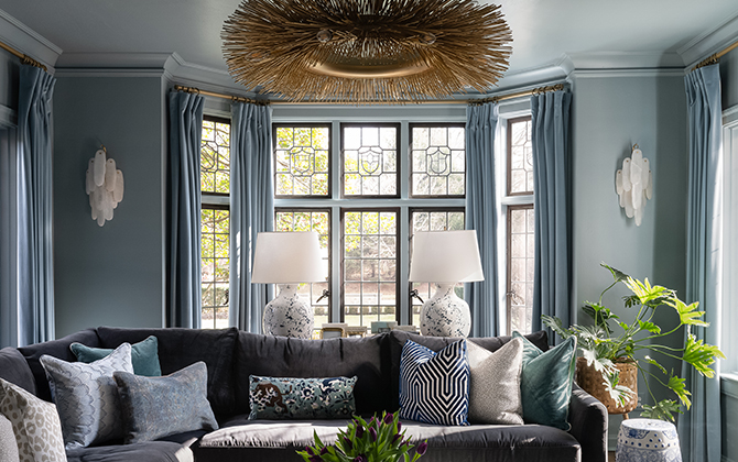 Living room with a dark gray sectional sofa, assorted pillows, marble coffee table with greenery, and tall windows with blue curtains.