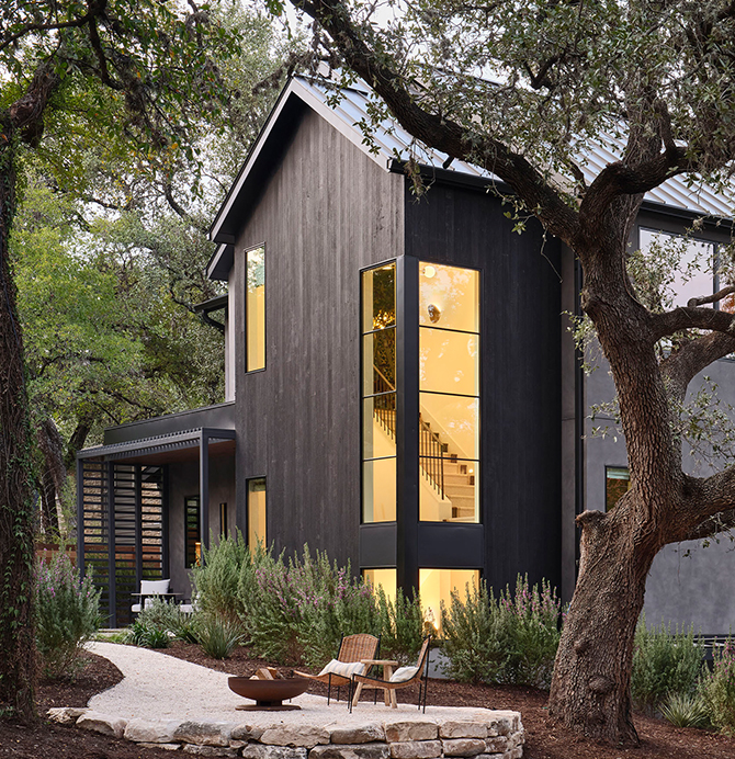 Modern home exterior with tall windows showing the staircase inside and a firepit with two chairs on a stone patio in the foreground.