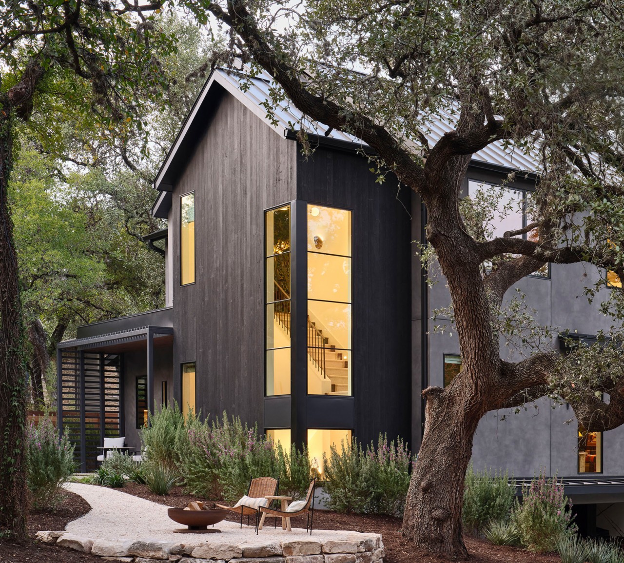 Modern home exterior with tall windows showing the staircase inside and a firepit with two chairs on a stone patio in the foreground.