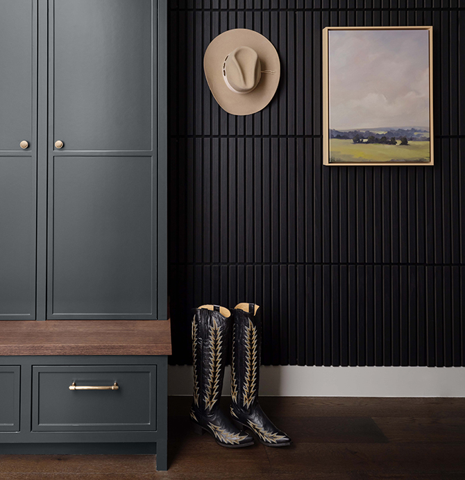 Mudroom featuring Rookwood Shutter Green cabinets with gold handles, black slatted wall, mounted cowboy hat, landscape painting, and patterned cowboy boots on wooden floor.