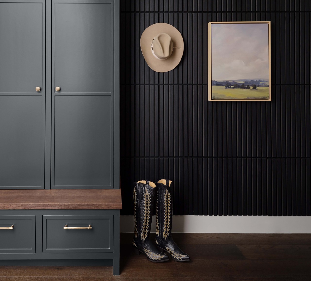 Mudroom featuring Rookwood Shutter Green cabinets with gold handles, black slatted wall, mounted cowboy hat, landscape painting, and patterned cowboy boots on wooden floor.