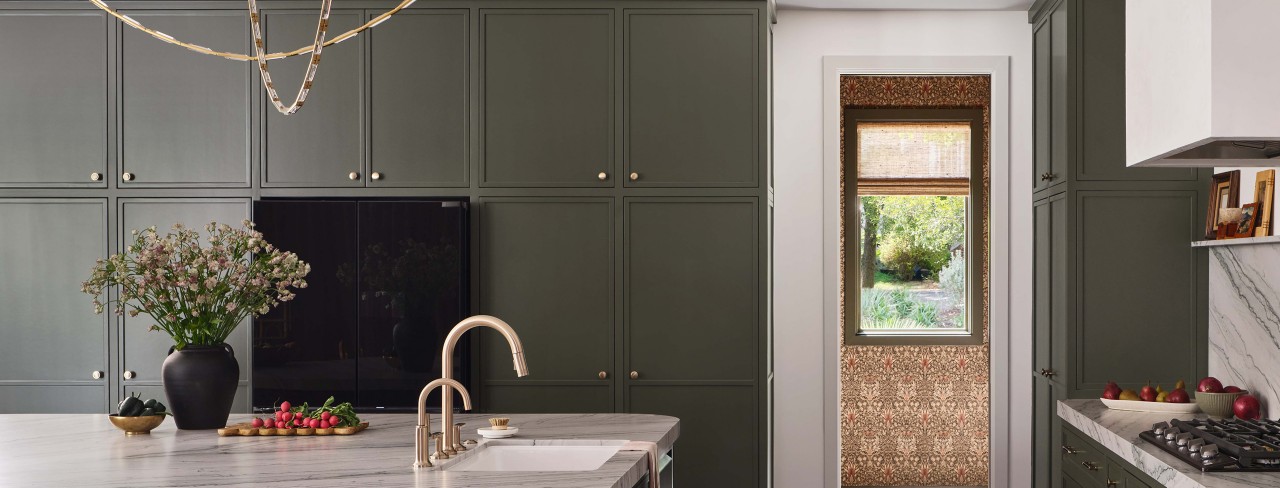 Modern kitchen with dark Roycroft Bronze Green cabinets, marble countertop, gold faucet, and a window view of a garden.