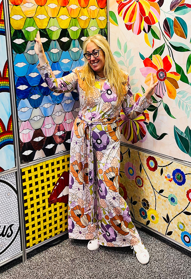 Allison Eden, wearing glasses and a patterned jumpsuit and posing in front of a collection of brightly colored tile mosaics.