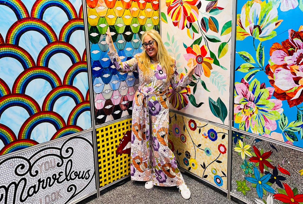 Allison Eden, wearing glasses and a patterned jumpsuit and posing in front of a collection of brightly colored tile mosaics.