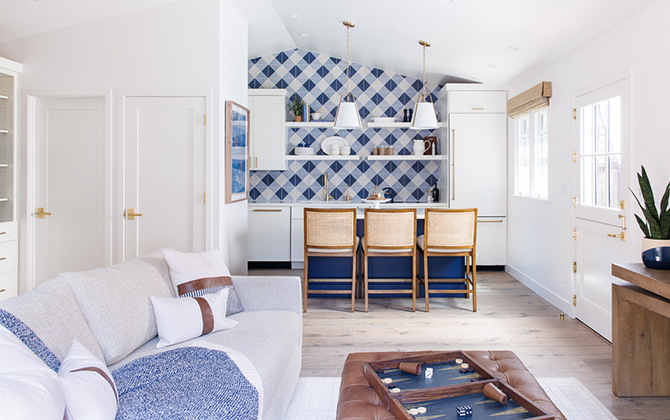 Modern living space with a light-colored couch, a tufted leather ottoman, and a kitchen area with a blue-and-white gingham patterned backsplash.