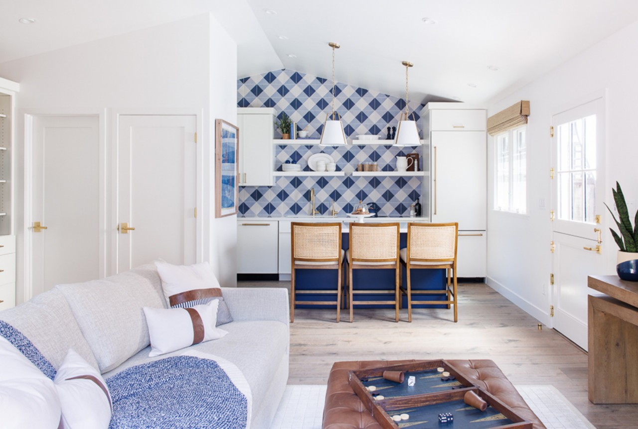 Modern living space with a light-colored couch, a tufted leather ottoman, and a kitchen area with a blue-and-white gingham patterned backsplash.