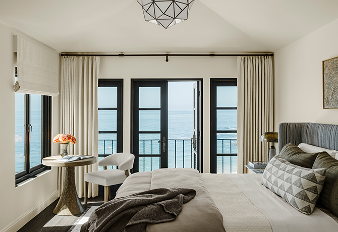 A Cheviot-painted bedroom with a view of the ocean through large black-framed windows, furnished with a bed, wooden table, and a chair.