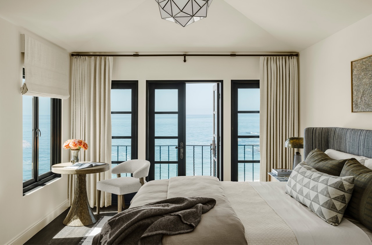 A Cheviot-painted bedroom with a view of the ocean through large black-framed windows, furnished with a bed, wooden table, and a chair.