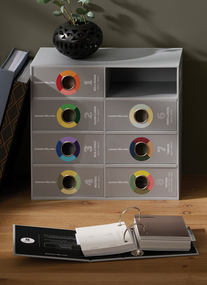 Gray box of color sample binders, small ringed binder of samples, and color fan deck on a wooden desktop with fabric samples and photos on a pinboard and a potted plant.