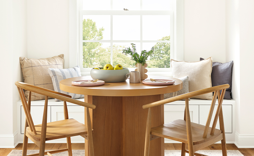 Naturally lit dining nook area with wood table and chairs in front of a window bench seat.