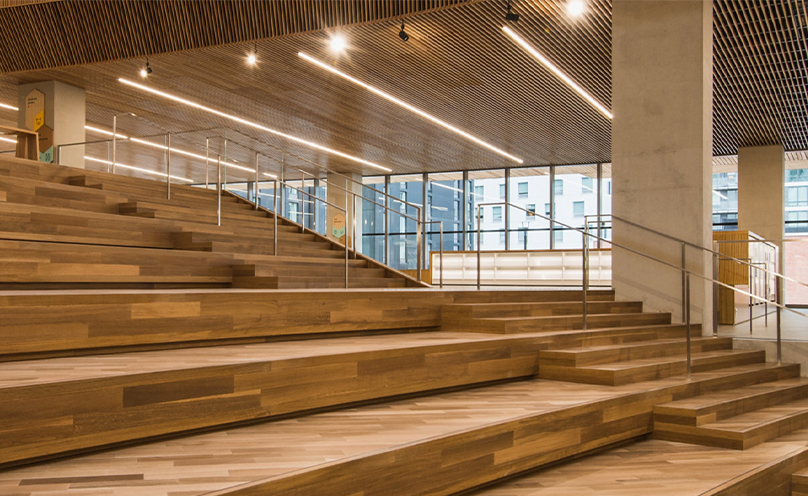 Wide wooden stairway inside a modern office space with stair steps at right and tiered stadium bench seating at left.