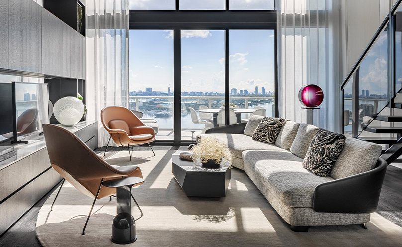 Modern urban loft living area with postmodern sectional and twin leather armchairs in front of flat screen TV with a view to an outside balcony area overlooking a city skyline.