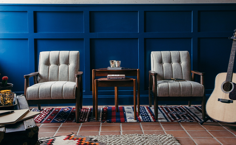 Two gray tufted chairs with wood frames and a shared accent table situated on woven rugs in front of a board-and-batten wall in a rich blue color, with an acoustic guitar on a stand off to the side.