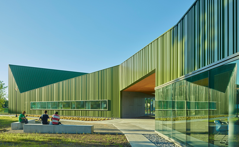 Exterior of Thaden school Reels and Wheels building in brilliant green metal with undulating roof in front of cloudless blue sky, students seated on cement benches in courtyard.