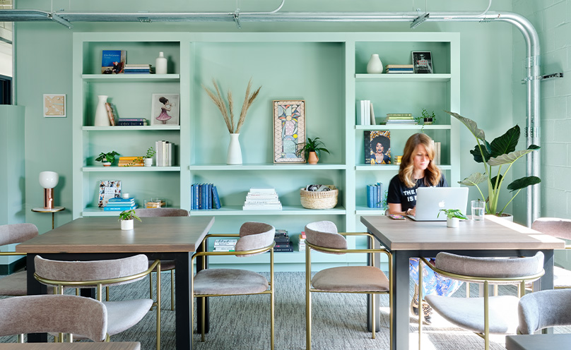 Interior of the Lola coworking space, with light blue-green walls and built-in shelving full of books and decor, with a woman working on a laptop at one of two modern worktables in foreground.