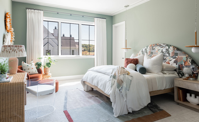 Soft green bedroom on a sunny day with lots of throw pillows and blankets on bed with oversized arch fabric-covered headboard, neutral accents and white curtains.