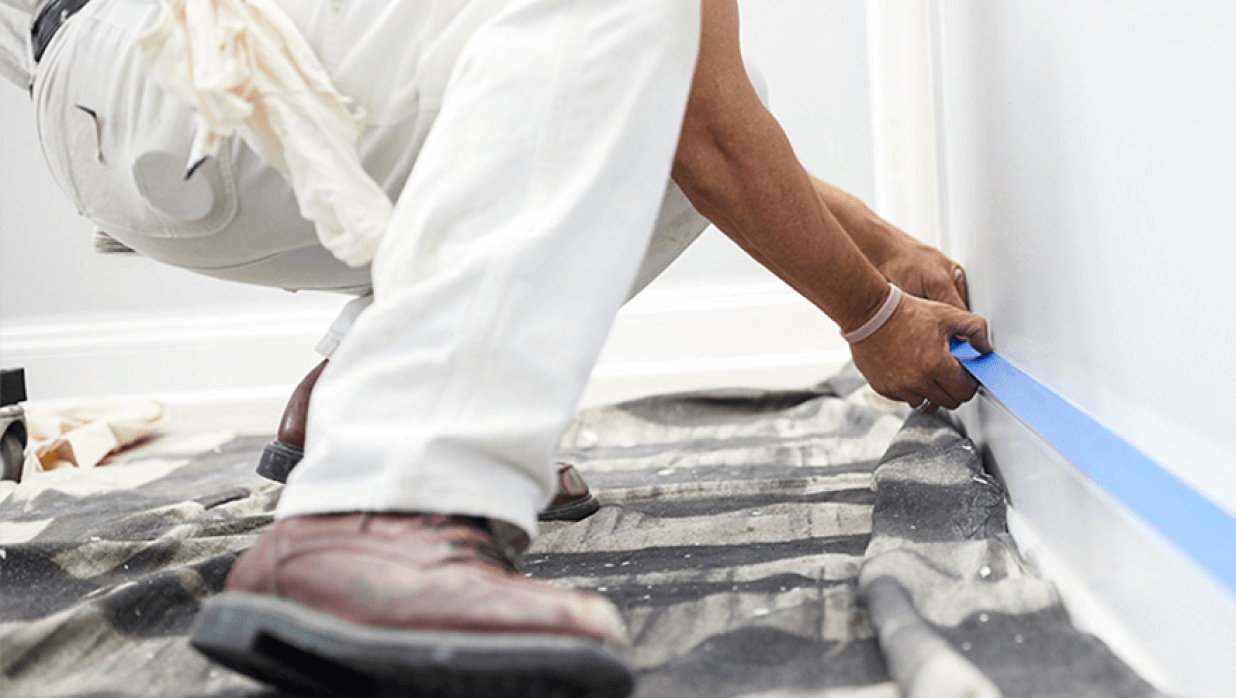A paint pro putting blue painter's tape along the top of a baseboard.