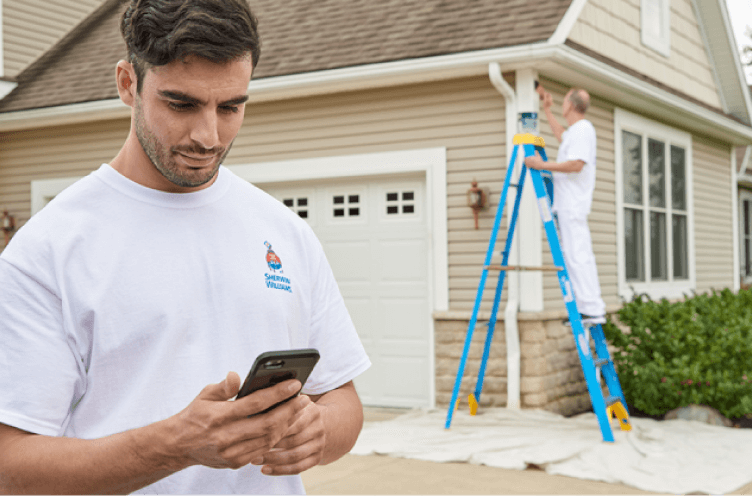 A contractor looking at his smart phone in a driveway of a house being painted by another contractor.