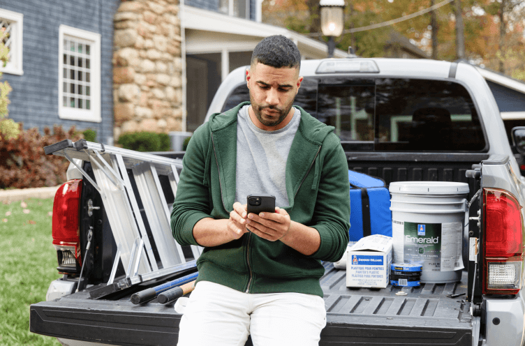 A paint pro sitting on the tailgate of a truck looking at a smartphone.