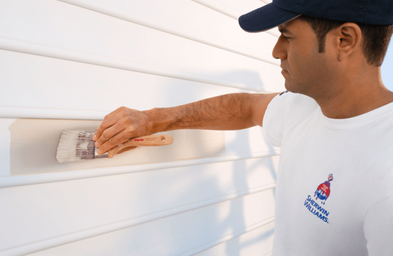 A person in a white shirt with the Sherwin-Williams logo painting exterior siding.