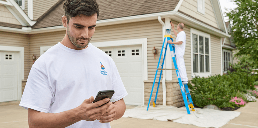 A contractor looking at his smart phone in a driveway of a house being painted by another contractor.