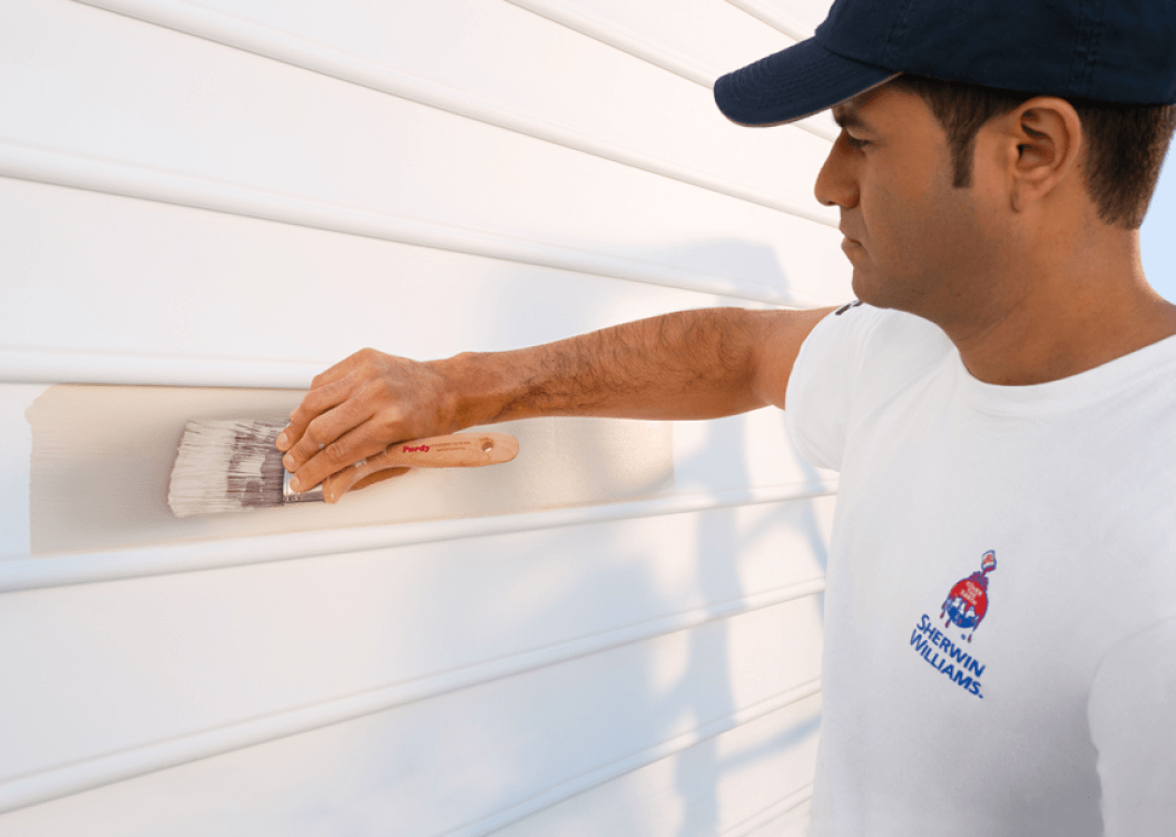 A person wearing a white shirt with a Sherwin-Williams logo painting vinyl siding.