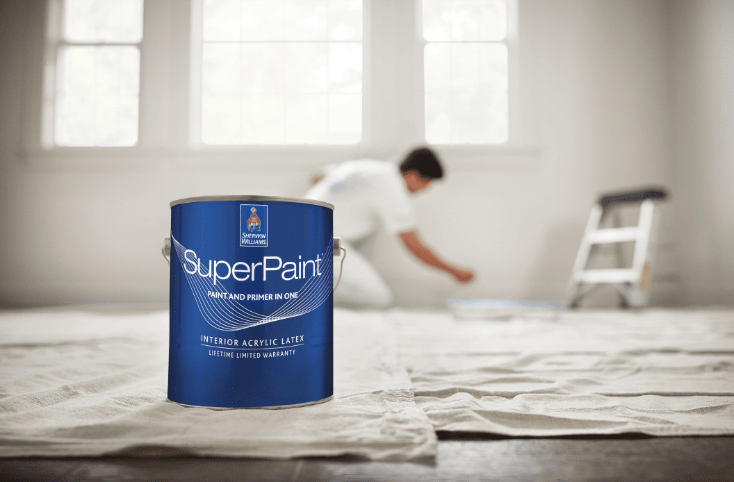 A can of SuperPaint on the floor near a person painting a wall.
