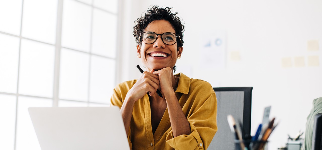Happy graphics designer sitting at her desk thinking about a design. Business woman smiling as she contemplates an idea. Creative woman working with a laptop and a stylus pen in an office.