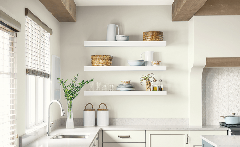 White kitchen with windows above the sink, wooden beams, and 3 floating shelves with kitchen items.