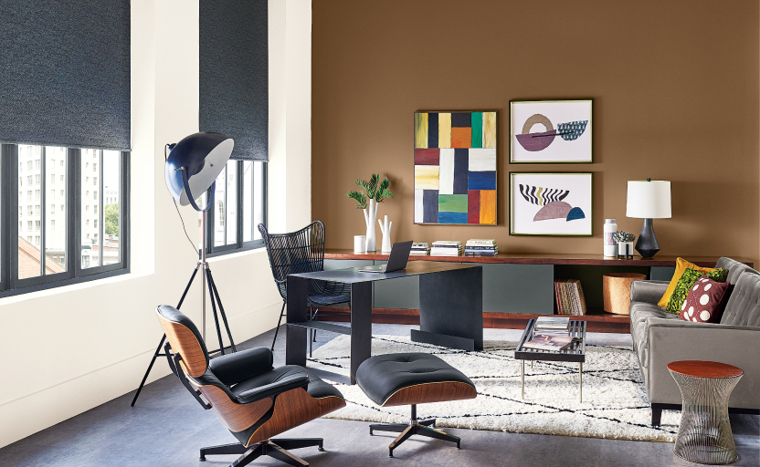 Modern office with one brown wall, one white wall, 2 windows, colorful art pieces, and neutral furnture.
