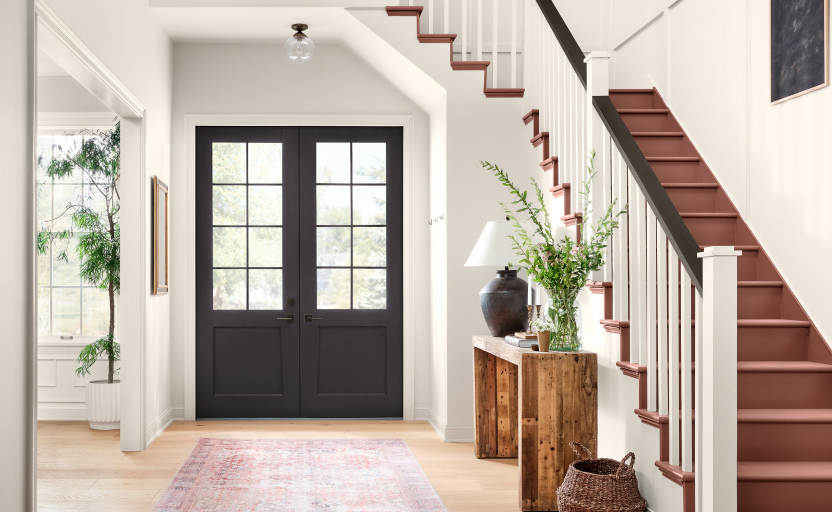 Well lit entryway with white walls, black door with windows, table with decor, and brown stairs.