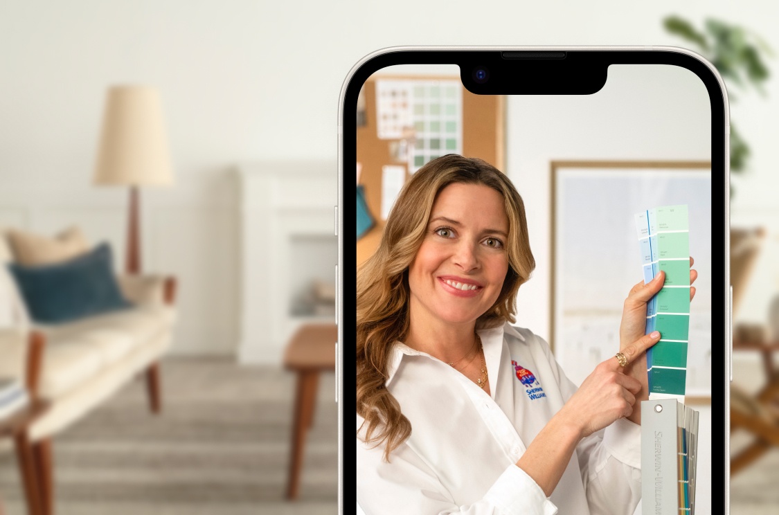 A phone showing a virtual color consultation with a woman holding up a fan deck. A white kitchen is blurred out behind the phone.